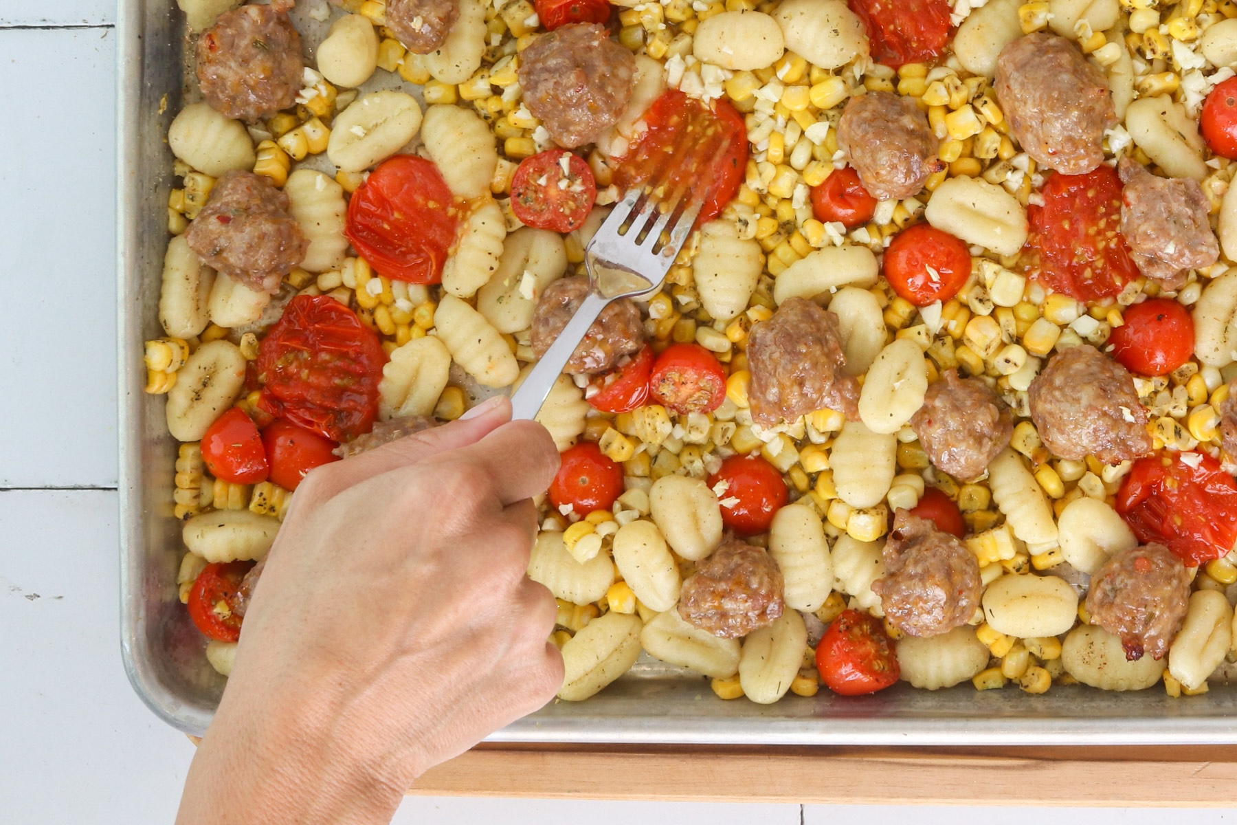 Sheet pan gnocchi with sausage, corn and roasted tomatoes being smashed with a fork to release their juices.