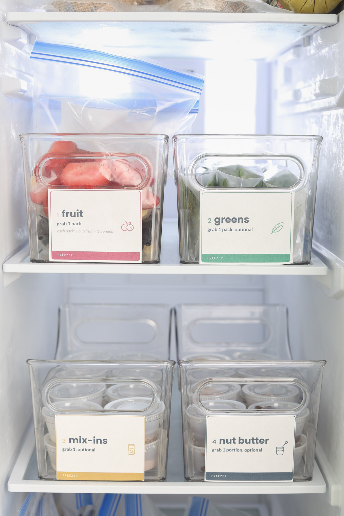 Labeled freezer bins organized for a make-your-own smoothie bar with fruit packs, spinach, mix-ins and portioned nut butter.
