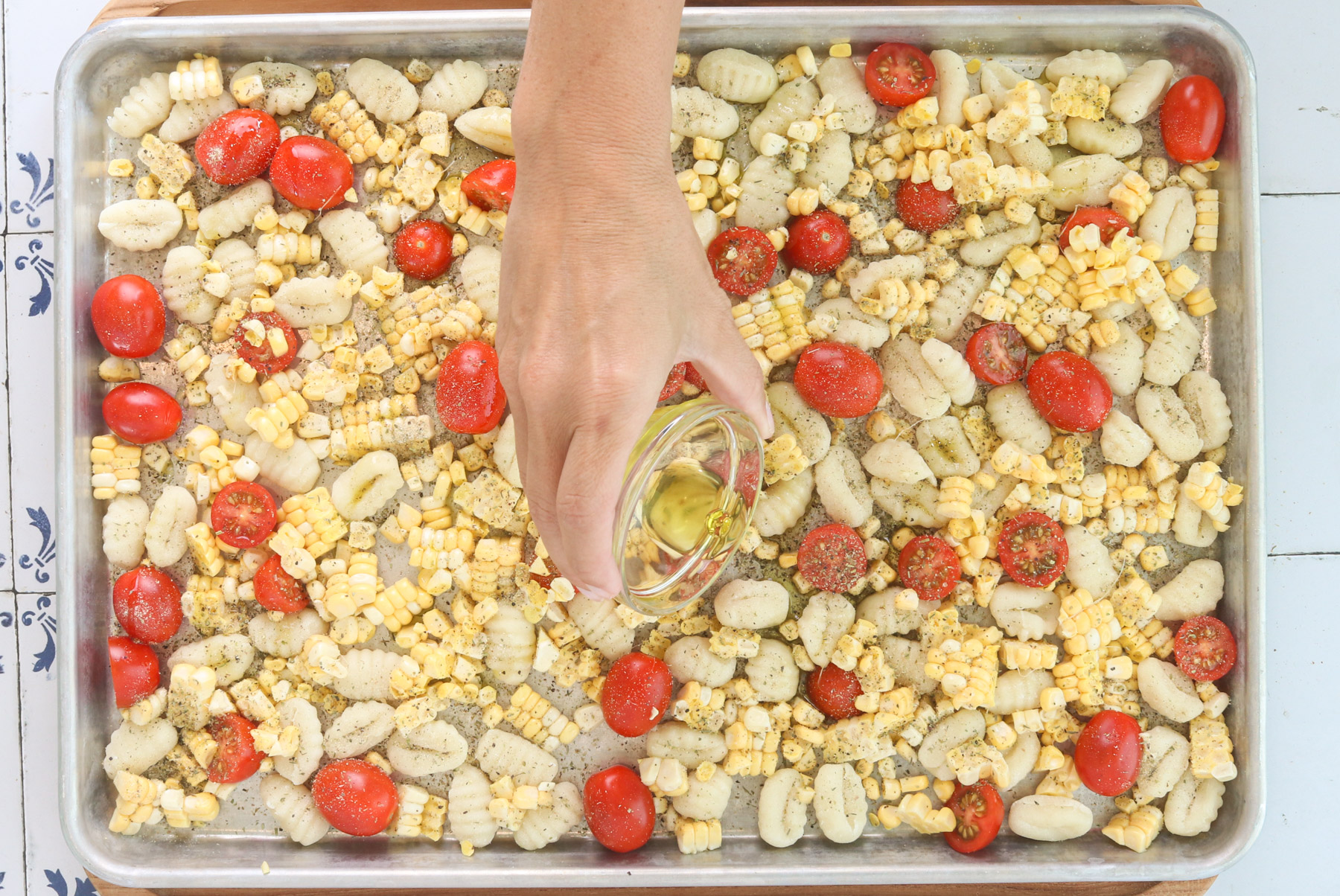 Olive oil being poured over gnocchi, corn and tomatoes on a sheet pan before roasting.