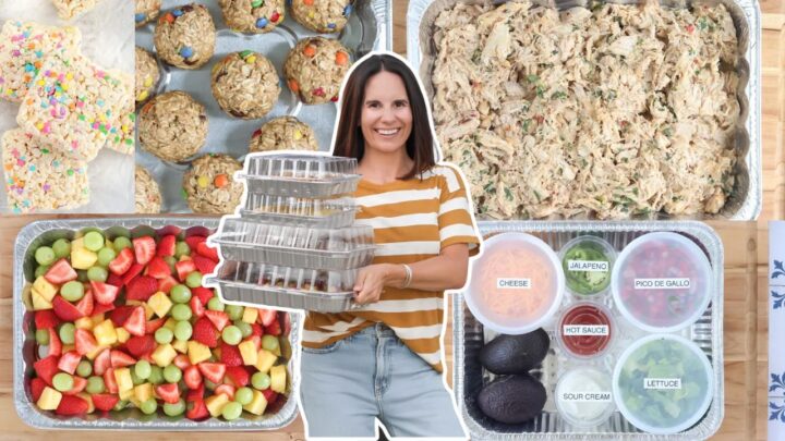 Woman holding stacked meal-prep containers surrounded by trays of shredded chicken, fresh fruit, rice cereal treats, energy bites, and taco toppings.