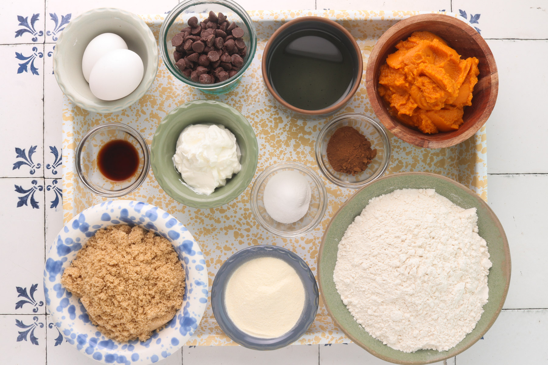 Ingredients for pumpkin protein muffins, including pumpkin puree, flour, vanilla protein powder, Greek yogurt, brown sugar, eggs, chocolate chips, oil, vanilla and spices arranged on a tray.