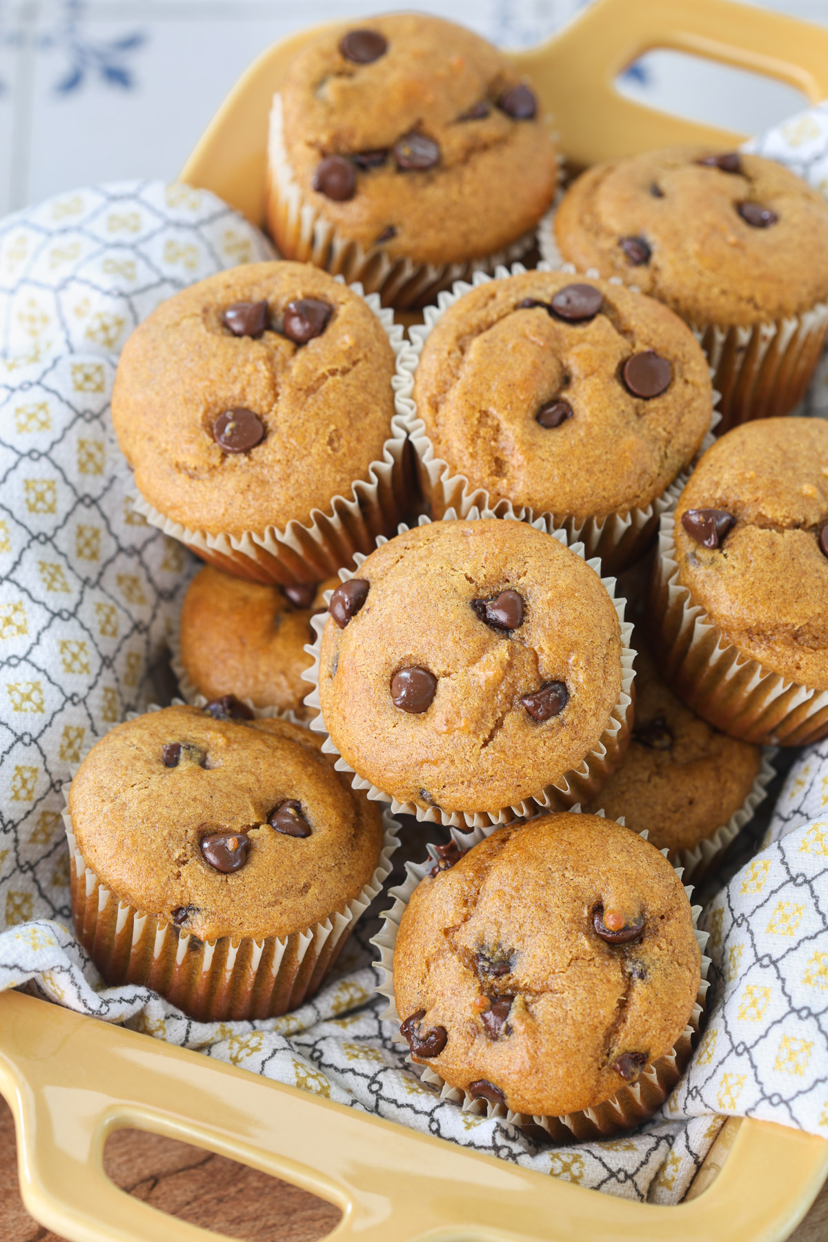 Pumpkin protein muffins with chocolate chips arranged in a yellow baking dish lined with a patterned kitchen towel.