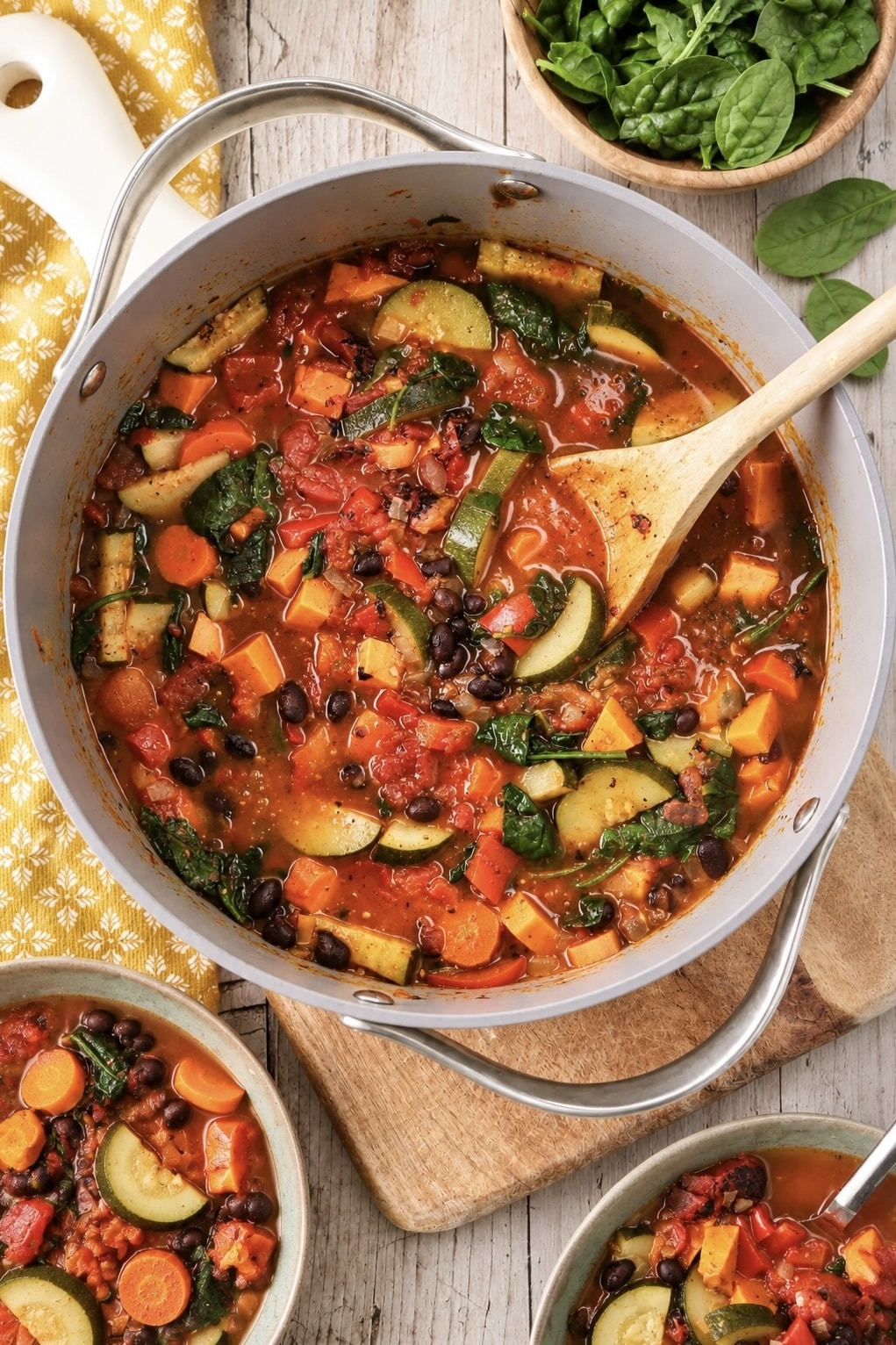 Overhead view of a large pot of chunky vegetable soup filled with zucchini, carrots, sweet potatoes, black beans, tomatoes, and spinach in a tomato-based broth, with a wooden spoon and bowls of soup nearby.